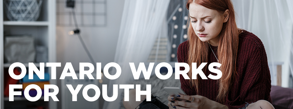 A young woman sits on her bed while looking at her smartphone.